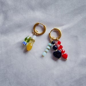 Puede incluir: Dos pendientes de aro dorados con abalorios de cuentas de colores. Un pendiente tiene un abalorio amarillo, verde, azul y blanco. El otro pendiente tiene un abalorio en forma de coraz&oacute;n azul, rojo y negro.