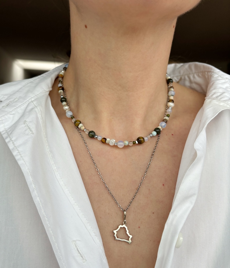 May include: A silver chain necklace with a pendant shaped like the outline of a state or country. The necklace is layered over a beaded necklace with white, brown, and green beads.