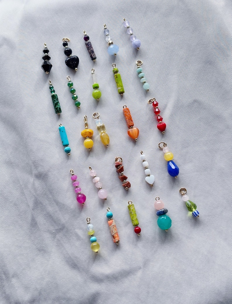 Puede incluir: Surtido de abalorios de piedras preciosas y vidrio de colores, cada uno con un lazo dorado para sujetar a joyas u otras manualidades. Los abalorios est&aacute;n dispuestos en un patr&oacute;n de arco&iacute;ris, con colores que incluyen rojo, naranja, amarillo, verde, azul, morado y rosa.