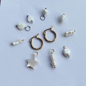 Puede incluir: Una colección de componentes de joyería que incluye dos pendientes de aro dorados y varios colgantes blancos. Los colgantes presentan formas como una estrella, un corazón, un círculo y cuentas rectangulares, hechos de materiales nacarados con anillas metálicas.