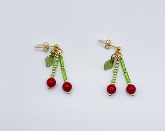 May include: A pair of gold cherry earrings with red and green beads. The earrings have a dangling design with two red beads and a green leaf on each earring.
