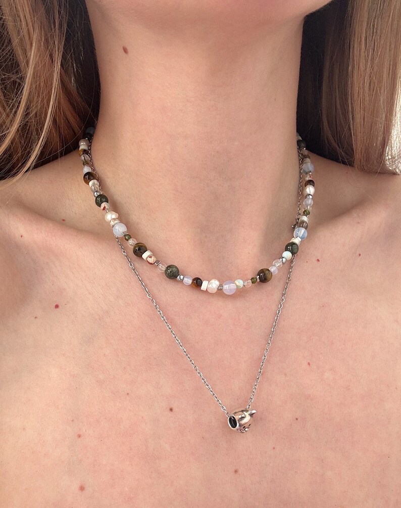 May include: A layered necklace with a silver chain and a small silver charm. The top necklace is made of various colored beads, including brown, green, white, and blue.