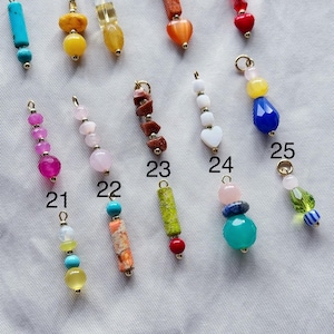 Puede incluir: Surtido de colgantes de joyer&iacute;a con cuentas de colores, cada uno con un dise&ntilde;o &uacute;nico. Los colgantes est&aacute;n numerados del 21 al 25 y presentan diversas formas, colores y materiales.