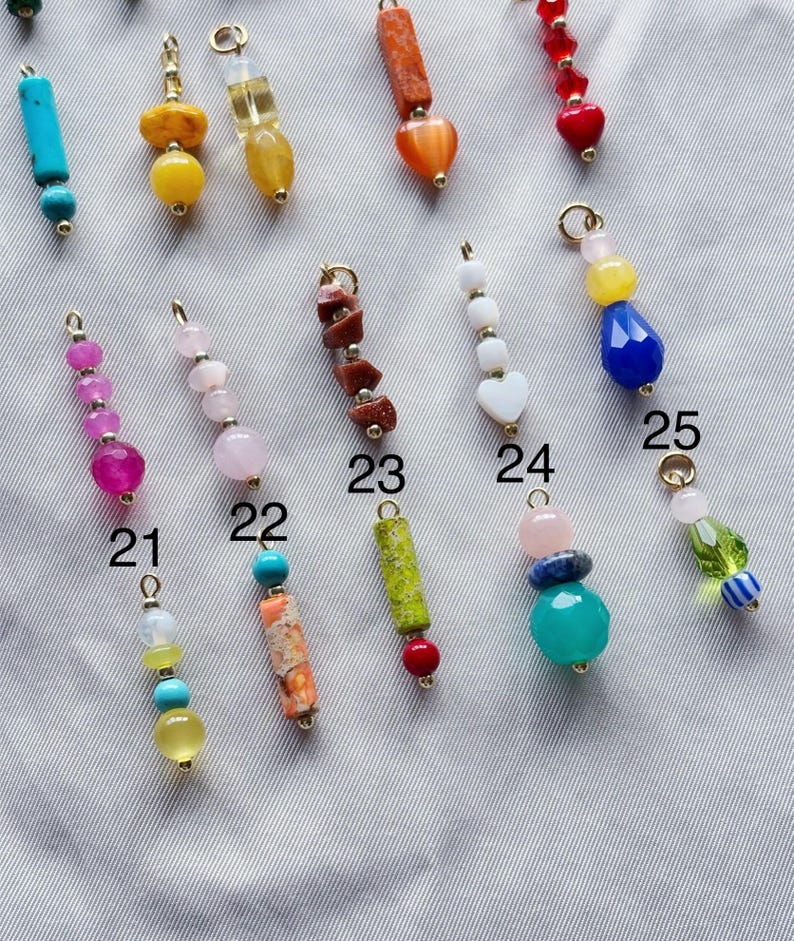 Puede incluir: Surtido de colgantes de joyer&iacute;a con cuentas de colores, cada uno con un dise&ntilde;o &uacute;nico. Los colgantes est&aacute;n numerados del 21 al 25 y presentan diversas formas, colores y materiales.
