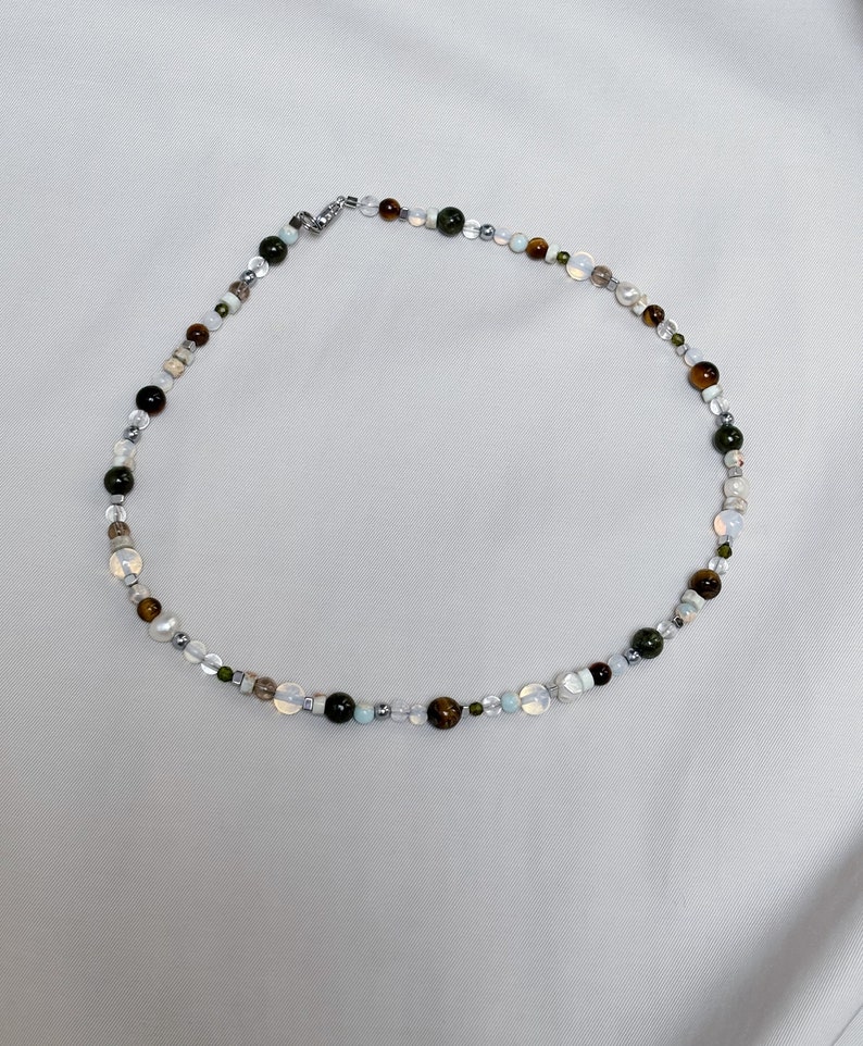 May include: A multi-colored beaded necklace with green, brown, white, and gray beads. The necklace is made of natural stones and has a silver clasp.