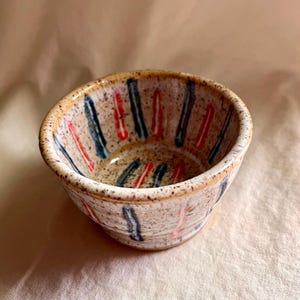 May include: A small, handmade ceramic bowl with a speckled beige exterior. The bowl features a series of vertical stripes in red and blue. The interior is a lighter beige color. The bowl is ideal for serving dips, snacks, or small portions.