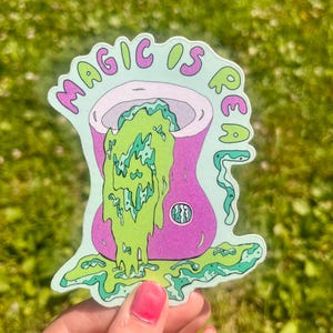 May include: A die-cut sticker featuring a purple cauldron with a light green, viscous liquid overflowing from the top and pooling at the base. The text MAGIC IS REAL is printed in purple and green letters arched above the cauldron.