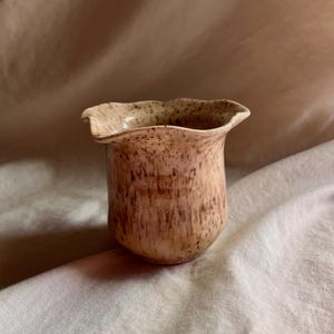 May include: A small, speckled ceramic vase with a flared, wavy rim. The vase has a light beige base color with brown speckles and vertical streaks. The interior is a lighter beige. The vase is likely handmade and suitable for small floral arrangements.