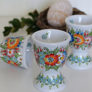 Op de afbeelding: Witte keramische eierdopjes met kleurrijke bloemmotieven in rood, oranje, blauw en groen. De eierdopjes hebben een voet en zijn versierd met gedetailleerde bloempatronen. Perfect voor Pasen.
