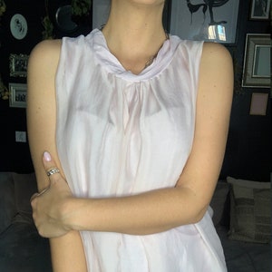 May include: A sleeveless, light pink blouse with a high, gathered neckline. The blouse is made of a sheer fabric and has a flowing, A-line silhouette.