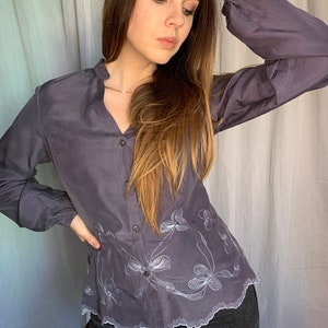 May include: A long-sleeved, gray button-down shirt with white embroidered floral designs. The shirt has a scalloped hem and a relaxed fit.