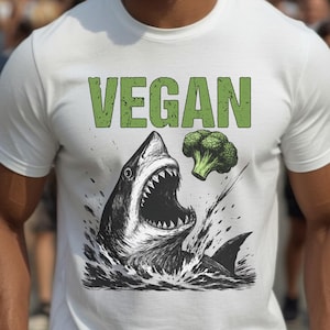May include: White t-shirt with the word "VEGAN" in green block letters above a black and white illustration of a shark with an open mouth and a broccoli floret. The shark is emerging from water.
