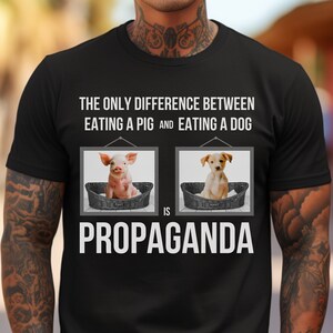 Op de afbeelding: Een zwart T-shirt met de tekst "Het enige verschil tussen het eten van een varken en het eten van een hond is propaganda." De tekst is wit en wordt omringd door twee afbeeldingen. De afbeelding links toont een varken in een mand en de afbeelding rechts toont een hond in een mand.
