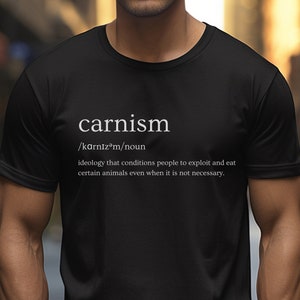 Puede incluir: Camiseta negra de cuello redondo con la palabra "carnism" en blanco, seguida de su pronunciación y definición. La definición explica el carnismo como una ideología que condiciona a las personas a explotar y comer ciertos animales incluso cuando no es necesario.
