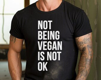 May include: Black crew neck t-shirt with the white text "NOT BEING VEGAN IS NOT OK". The shirt is made of a soft material and has short sleeves. The shirt is a statement piece for those who support veganism.