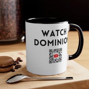 Peut inclure: Mug en céramique blanche avec intérieur et anse noirs. Le mug porte l'inscription "WATCH DOMINION" en noir, ainsi qu'un code QR menant à YouTube. Le mug est posé sur une surface en bois avec une cuillère, des grains de café et un macaron.