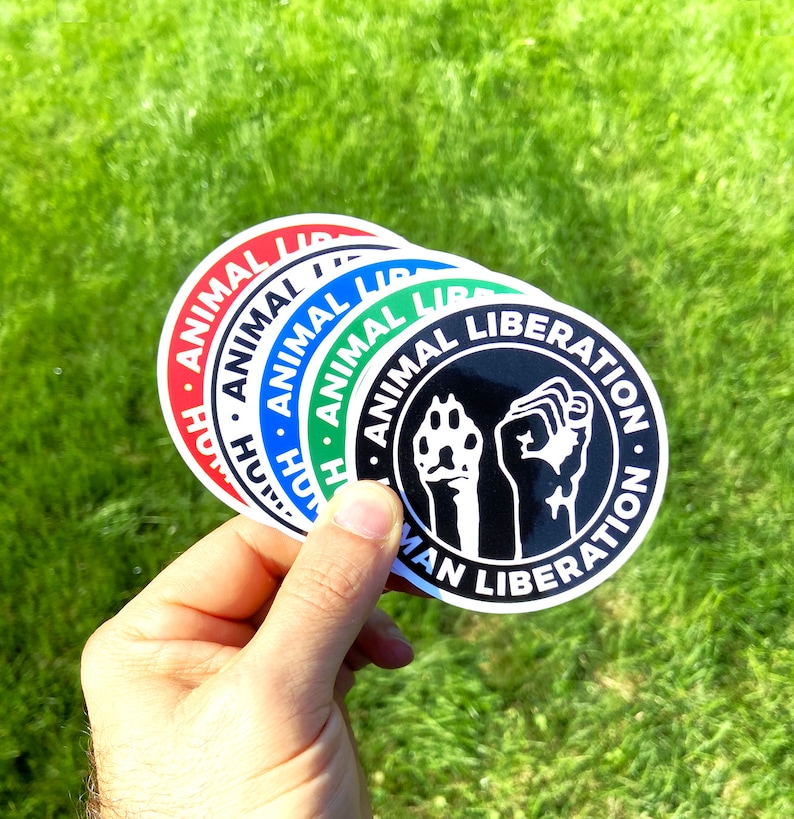 May include: Four round stickers with the text "Animal Liberation" and a black and white graphic of a fist and paw. The stickers are layered on top of each other, with the red sticker on the bottom, followed by the blue, green, and black stickers.
