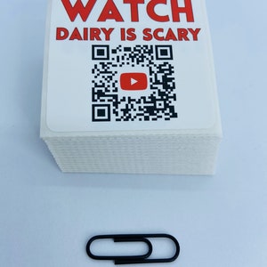Pode incluir: Uma pilha de adesivos quadrados brancos com texto vermelho que diz "WATCH DAIRY IS SCARY" e um c&oacute;digo QR preto e branco com um bot&atilde;o de reprodu&ccedil;&atilde;o vermelho no centro. Um clipe de papel preto est&aacute; no centro inferior da imagem.