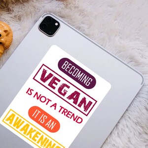 Op de afbeelding: Een witte sticker met de tekst "Vegan worden is geen trend, het is een ontwaken" in rood, paars en oranje. De tekst is in een vet, verweerd lettertype.
