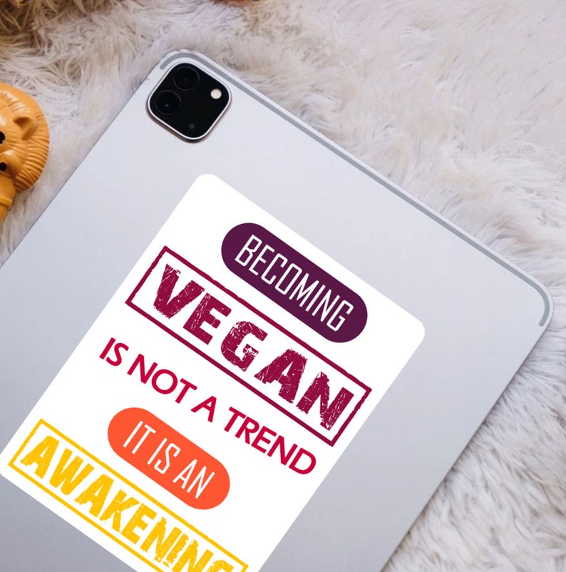 Op de afbeelding: Een witte sticker met de tekst "Vegan worden is geen trend, het is een ontwaken" in rood, paars en oranje. De tekst is in een vet, verweerd lettertype.