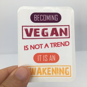 Op de afbeelding: Witte sticker met de tekst "Vegan worden is geen trend, het is een ontwaken" in rode, oranje en gele letters.