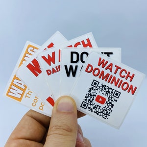 Pode incluir: Uma m&atilde;o segurando um monte de adesivos brancos com texto vermelho e preto. Os adesivos dizem "Watch Dominion", "Don't Watch", "Win", "Win", e "Win". Um adesivo tem um c&oacute;digo QR com um bot&atilde;o de reprodu&ccedil;&atilde;o vermelho no centro.