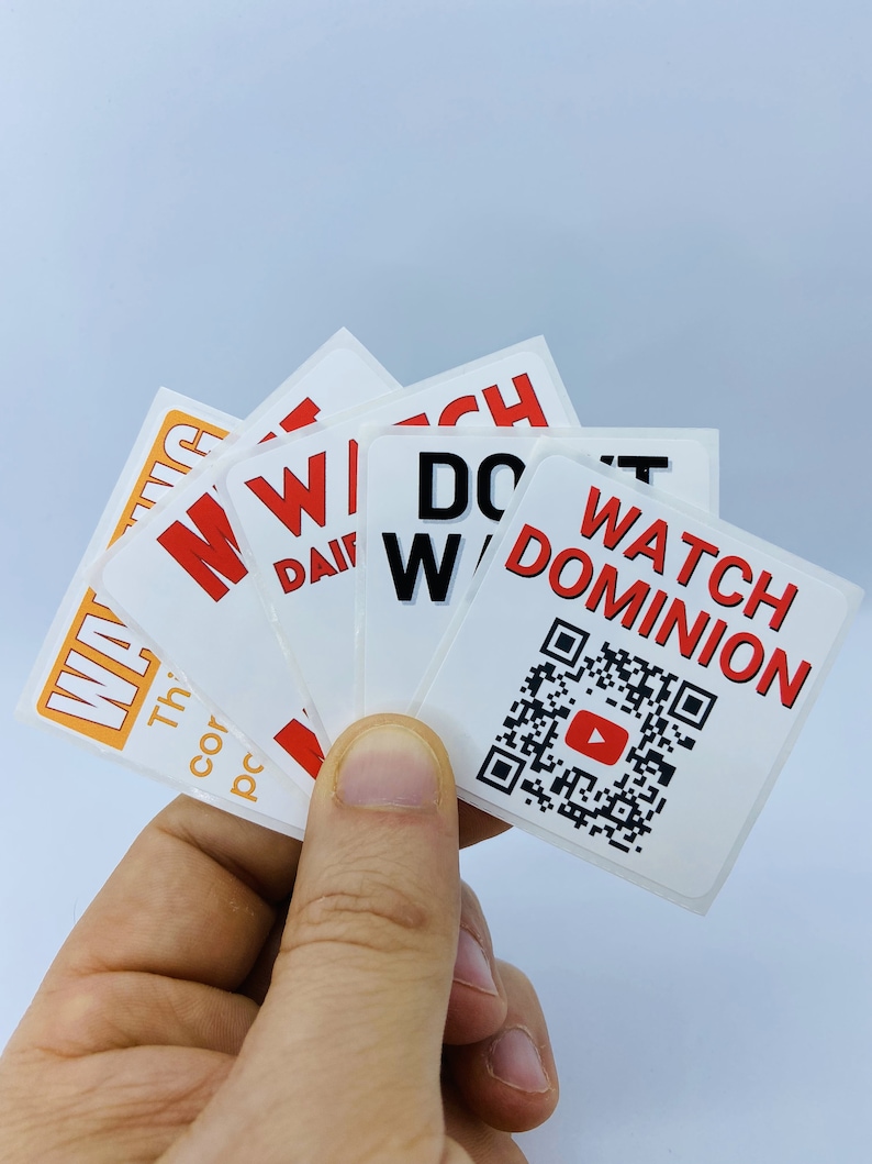 Pode incluir: Uma m&atilde;o segurando um monte de adesivos brancos com texto vermelho e preto. Os adesivos dizem "Watch Dominion", "Don't Watch", "Win", "Win", e "Win". Um adesivo tem um c&oacute;digo QR com um bot&atilde;o de reprodu&ccedil;&atilde;o vermelho no centro.