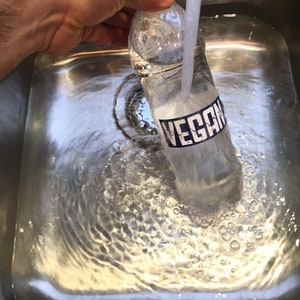 May include: A clear plastic water bottle with a blue label that says "VEGAN" is being rinsed under a faucet in a stainless steel sink.