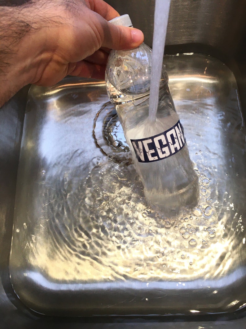May include: A clear plastic water bottle with a blue label that says "VEGAN" is being rinsed under a faucet in a stainless steel sink.