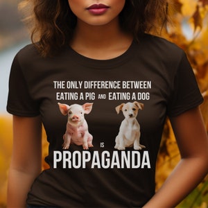 Op de afbeelding: Een bruin T-shirt met de tekst "Het enige verschil tussen het eten van een varken en het eten van een hond is propaganda."  Een varken en een hond zijn afgebeeld onder de tekst.