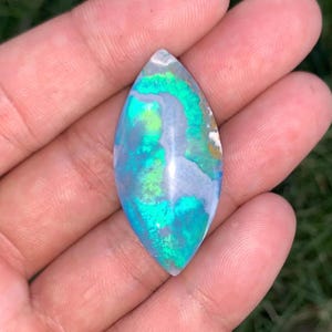 Peut inclure: Une pierre précieuse cabochon en forme de marquise présentant une surface lisse et polie avec des tourbillons vibrants de bleu iridescent, de vert vif et de gris doux. La pierre affiche un effet de jeu de couleurs sur toute sa surface.