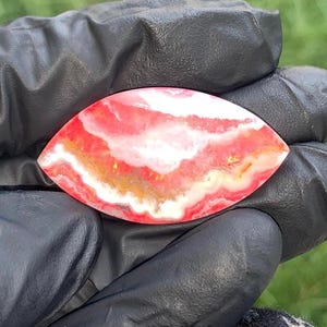May include: A marquise-shaped rhodochrosite gemstone cabochon featuring vibrant bands of pink, white, and soft orange. The stone displays a natural, wavy, layered pattern across its polished surface, showcasing distinct colour variations throughout.