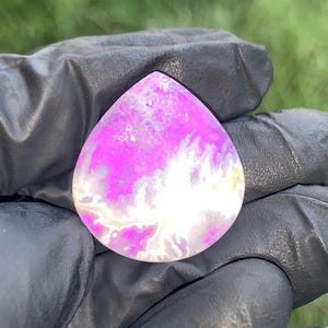 May include: A teardrop-shaped cabochon stone featuring a vibrant magenta and white marbled pattern with translucent areas. The polished surface displays organic, cloud-like inclusions and swirls of deep pink color throughout the gemstone.