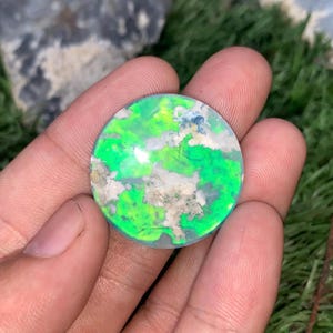 Può includere: Una gemma rotonda e lucidata con un vivace motivo verde e bianco. La pietra presenta un design a vortice, simile a una nuvola, con sfumature di blu e grigio. La gemma è tenuta in una mano, mettendo in risalto la sua bellezza naturale e le sue uniche variazioni di colore.