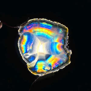 May include: A translucent, iridescent gemstone slice with a rainbow of colors, including yellow, orange, blue, and green. The stone has a rough, textured edge and a crystalline interior. The background is black.