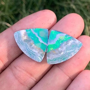 Peut inclure: Deux cabochons de forme triangulaire présentant un motif à bandes bleu sarcelle et blanc vif avec des éclats irisés verts et dorés. Les pierres ont une surface lisse et polie et présentent des couches ondulées distinctes de couleur.