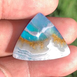 May include: A triangular-shaped gemstone featuring a smooth, polished surface with horizontal bands of translucent light blue, opaque white, and a textured, earthy golden-brown layer running through the center of the stone.