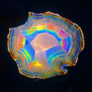 May include: A translucent, circular agate slice featuring concentric bands of vibrant rainbow colors including yellow, blue, and orange. The mineral specimen has a jagged, irregular outer edge with a metallic gold-toned rim against a black background.