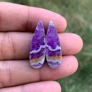 Peut inclure: Deux cabochons en forme de goutte d'eau présentant une teinte violette vibrante avec des bandes horizontales distinctes de blanc et de brun doré. Les pierres ont une surface lisse et polie et présentent un motif minéral naturel en couches.