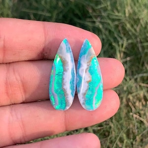 May include: Two pear-shaped cabochons featuring a white base with prominent, vibrant teal and green iridescent bands running vertically through the center. The stones have a smooth, polished surface and display a distinct color play pattern.