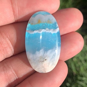 May include: An oval-shaped cabochon gemstone featuring a smooth, polished surface with horizontal bands of sky blue, translucent turquoise, and opaque white. The stone displays a layered, cloud-like pattern with subtle hints of golden-brown inclusions.
