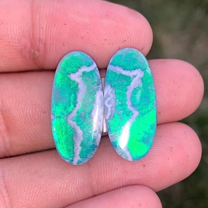 May include: Two oval-shaped cabochons featuring a vibrant green iridescent surface with a distinct, jagged white vein running vertically through the center of each stone, displaying a polished, smooth, and reflective finish on both pieces.