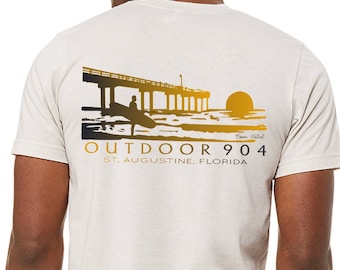 May include: A beige t-shirt with a graphic of a pier, a surfer, and a sunset. The text "OUTDOOR 904 ST. AUGUSTINE, FLORIDA" is printed on the shirt.