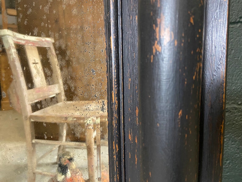 May include: A black wooden framed mirror with a distressed finish. The mirror has a slightly cloudy appearance and reflects a wooden chair.