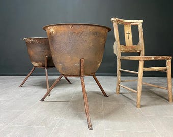 May include: Two large, weathered, round cast iron cauldrons with rusted surfaces, each supported by a three-legged metal stand. Beside them stands a wooden chair with a worn finish and a small cross symbol carved into the backrest.