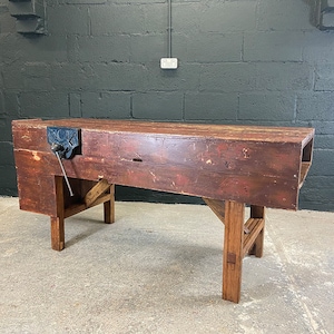 May include: A vintage wooden workbench with a metal vice and a dark brown finish. The workbench has a rectangular top and two wooden legs.
