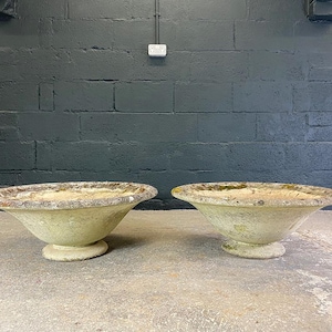 May include: A pair of weathered, off-white stone planters with wide, shallow basins and circular pedestal bases. The exterior surfaces show signs of age, including textured discolouration, mossy patches, and a rough, porous finish throughout.