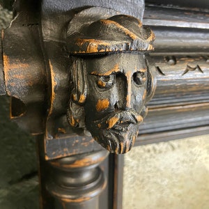 May include: A dark wood carved frame with a detailed, three-dimensional face of a man with a beard and a hat. The face is painted black and has a distressed look.