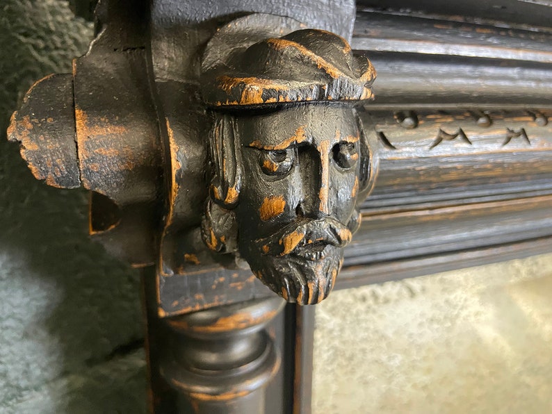 May include: A dark wood carved frame with a detailed, three-dimensional face of a man with a beard and a hat. The face is painted black and has a distressed look.