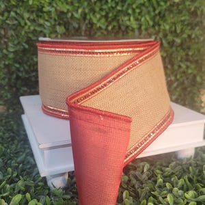 May include: A roll of tan burlap ribbon featuring a smooth, solid red fabric border on both edges. The ribbon is wound into a spool shape, showcasing the textured, woven tan center contrasted by the vibrant red trim and metallic gold accents.